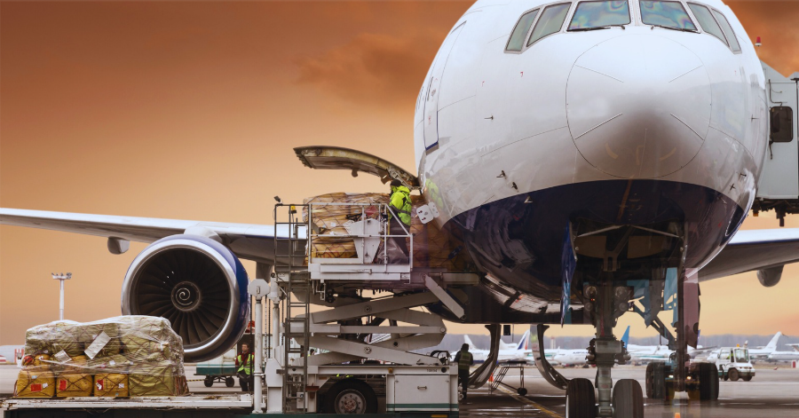 Cargo airplane with its forward cargo door open being loaded with palletized freight on an airport tarmac at sunset