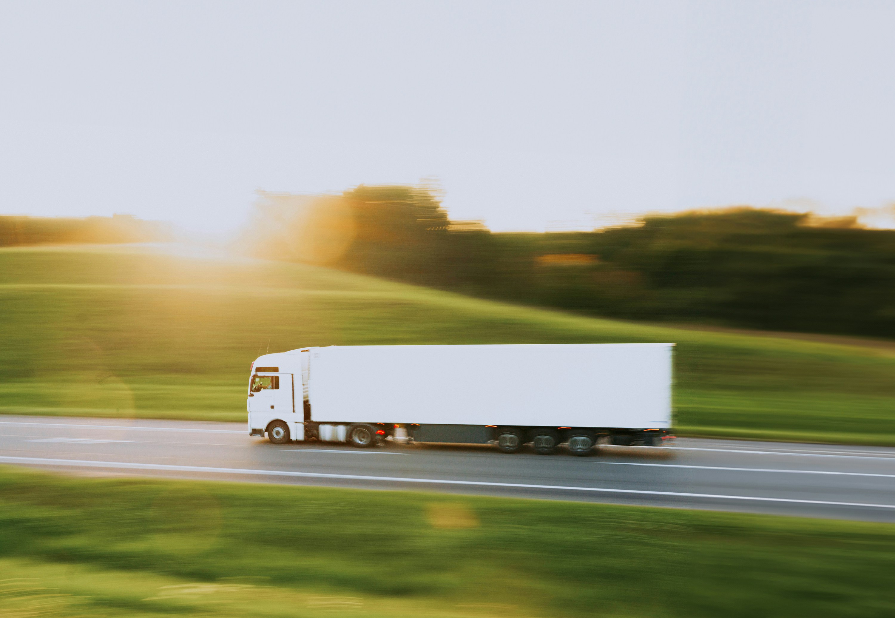 White semi truck speeding down a highway past green fields in warm sunlight
